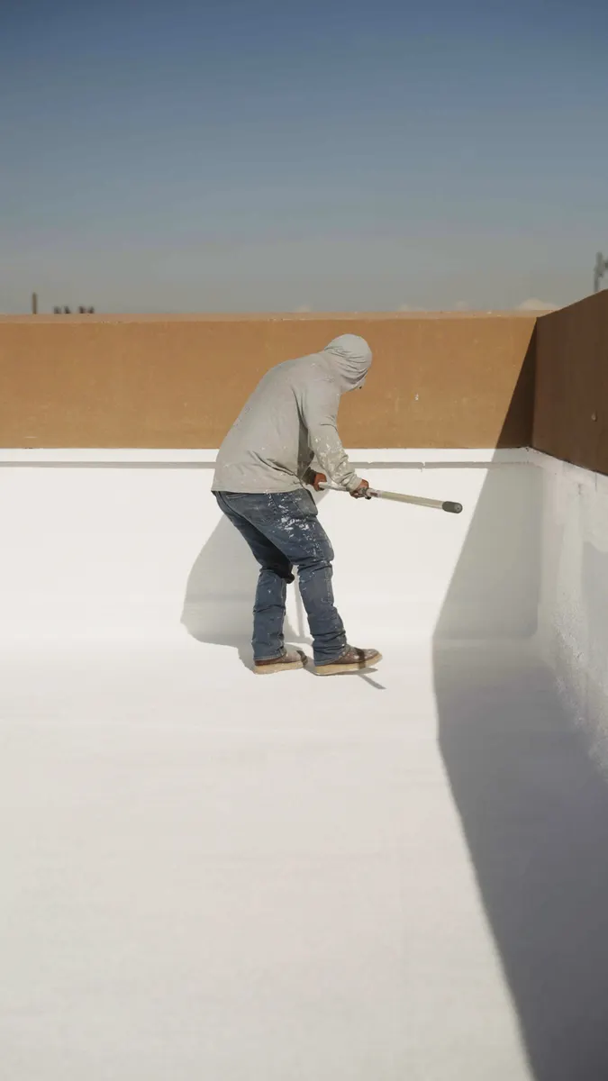 Roofer rolling white coating along the base of a parapet wall on a flat roof